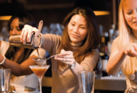 Un grupo de participantes en una clase de elaboración de cócteles se reunió alrededor de una estación de bar, mezclando ingredientes y preparando bebidas bajo la guía de un instructor.