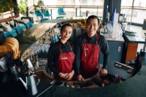 "Dos camareros con delantales sonriendo detrás de una barra en un restaurante moderno"